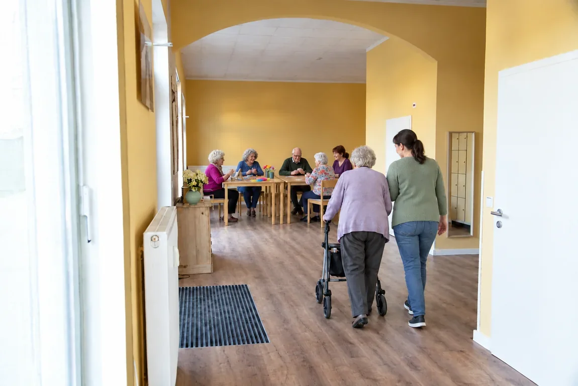 Eine Seniorin geht mit Rollator und Begleitung durch den Eingangsbereich, hinten basteln und spielen Gäste an Tischen